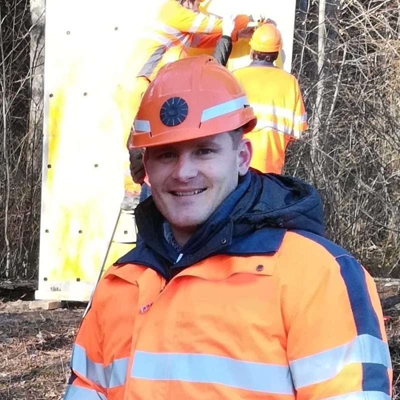 martin-ebneter-dynacem-schweiz-x-effects-gmbh Martin Ebneter mit orangem Helm und Arbeitskleidung. Im Hintergrund zwei Arbeiter bei der Vorbereitung einer Sprengung.